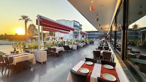 Un restaurante al aire libre con mesas y sillas, una piscina al fondo y un atardecer.