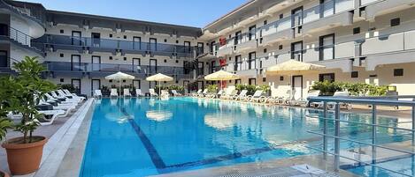 A swimming pool with lounge chairs and umbrellas, surrounded by a multi-story building with balconies.