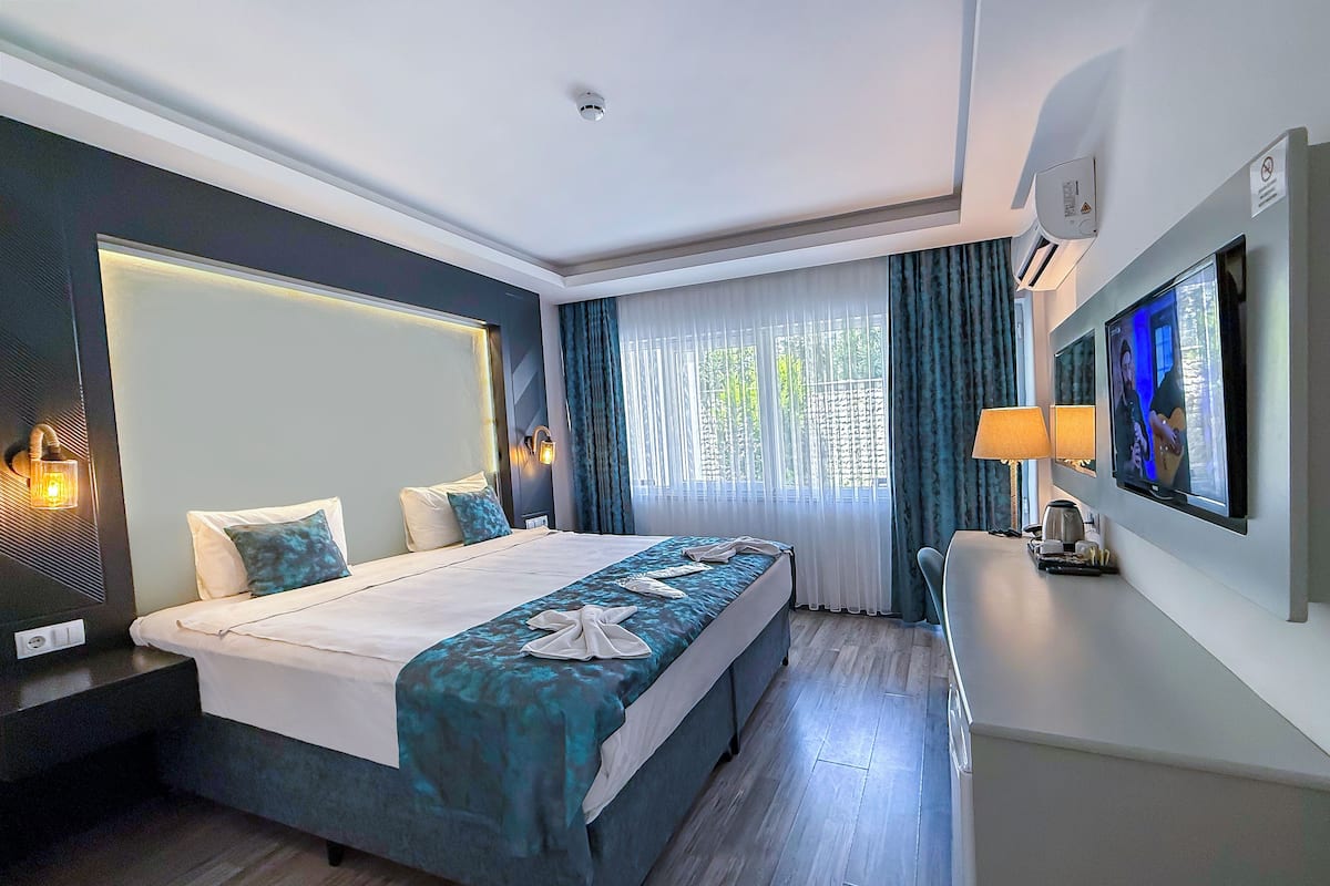 A modern hotel room with a large bed, a flat-screen TV, a desk with a lamp, and a view of greenery through the window.