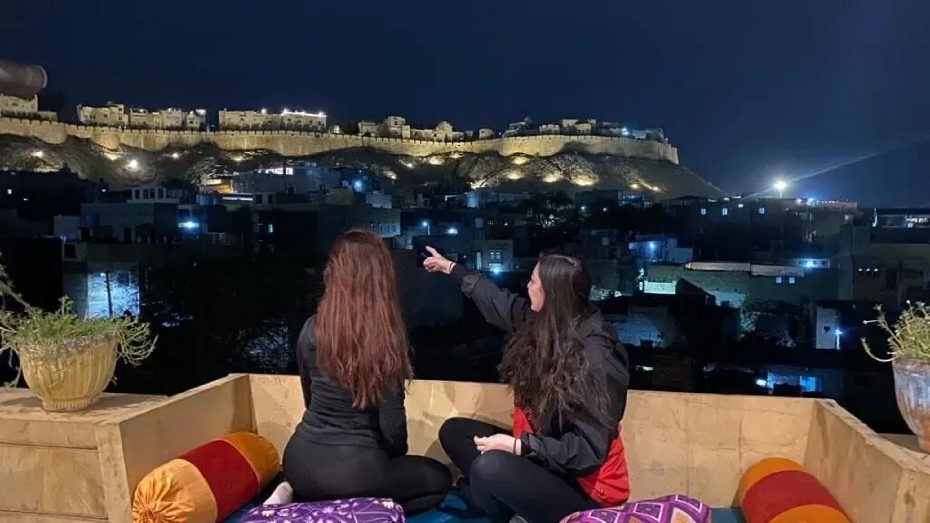 Two individuals seated on a balcony overlooking a historic fortified city with illuminated buildings.