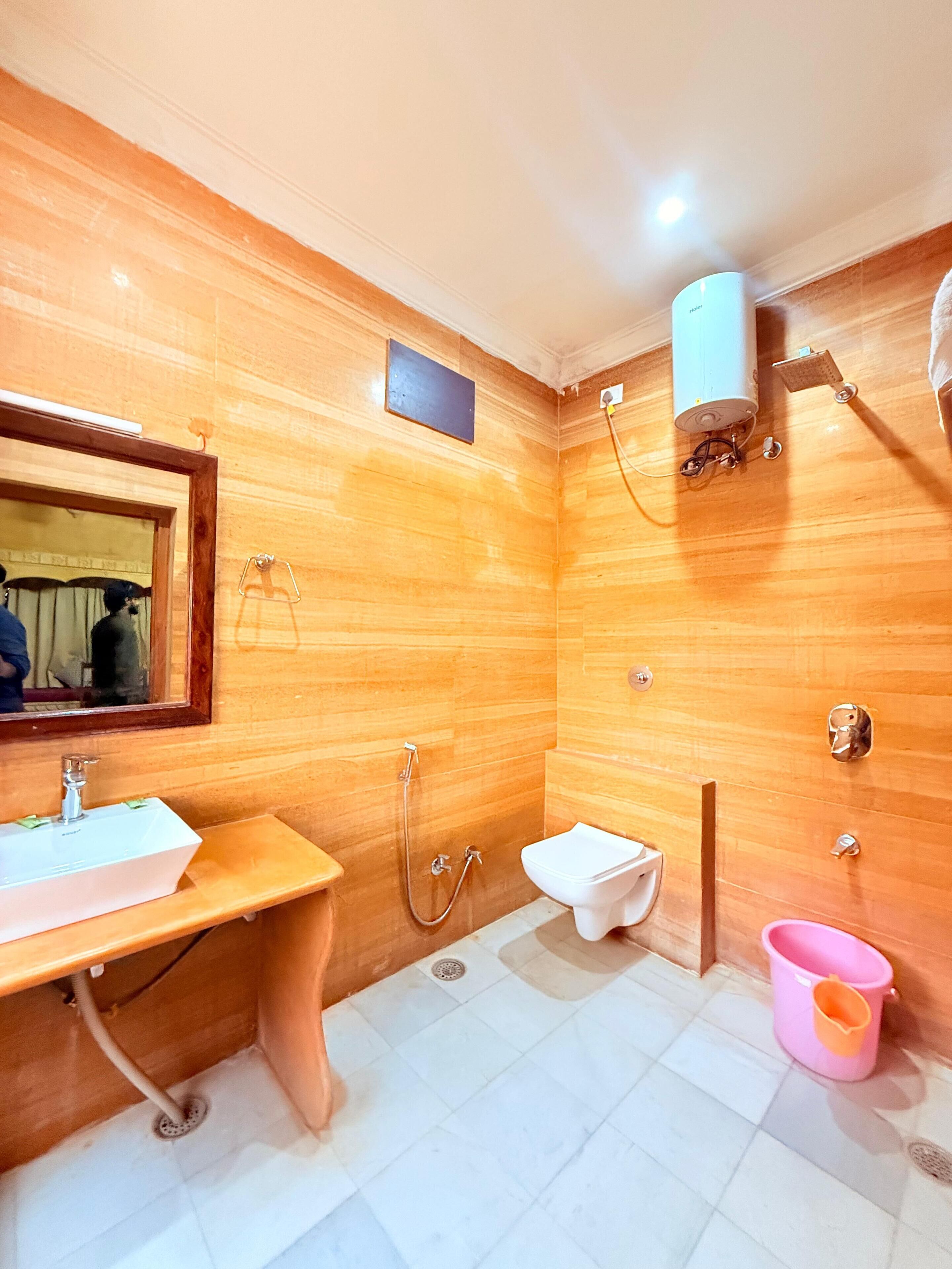 A bathroom with a wooden vanity, a sink, a mirror, a toilet, a shower, and a pink bucket.