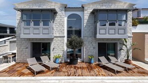 Un edificio de dos pisos con fachada de piedra, un área de piscina con sillones de descanso y un árbol en maceta.