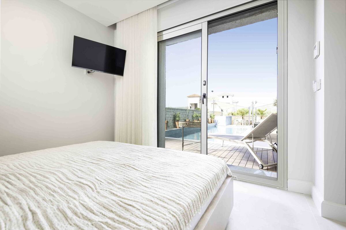 Une chambre avec un lit, un téléviseur fixé au mur et une porte-fenêtre coulissante donnant sur un balcon avec une piscine et du mobilier d’extérieur.