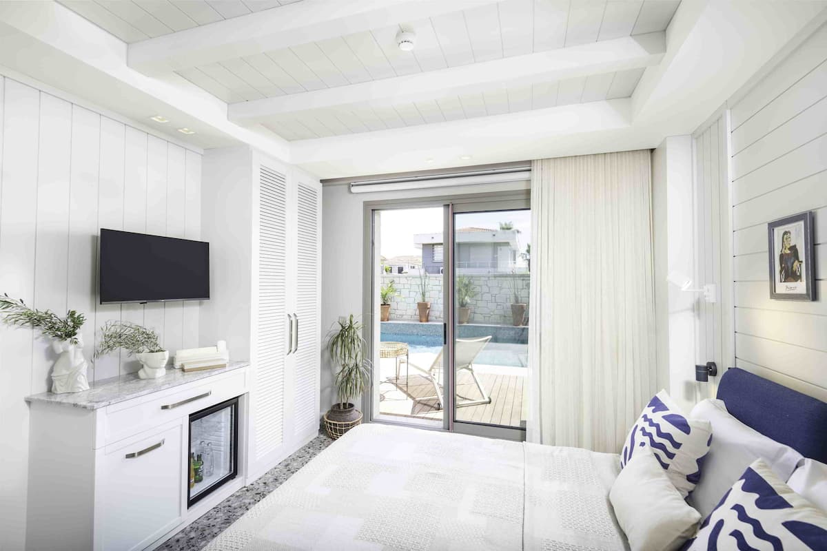 Une chambre moderne avec un grand lit, un téléviseur fixé au mur, une porte-fenêtre coulissante donnant sur un balcon et un petit meuble sur lequel est posé un autre téléviseur.
