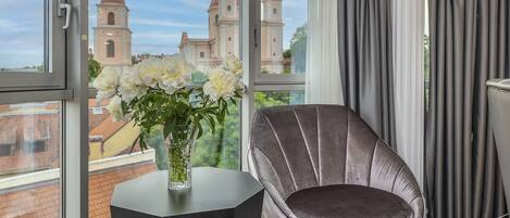 Una habitación con un ventanal grande, una silla, un florero con flores y vistas a un edificio con torres.