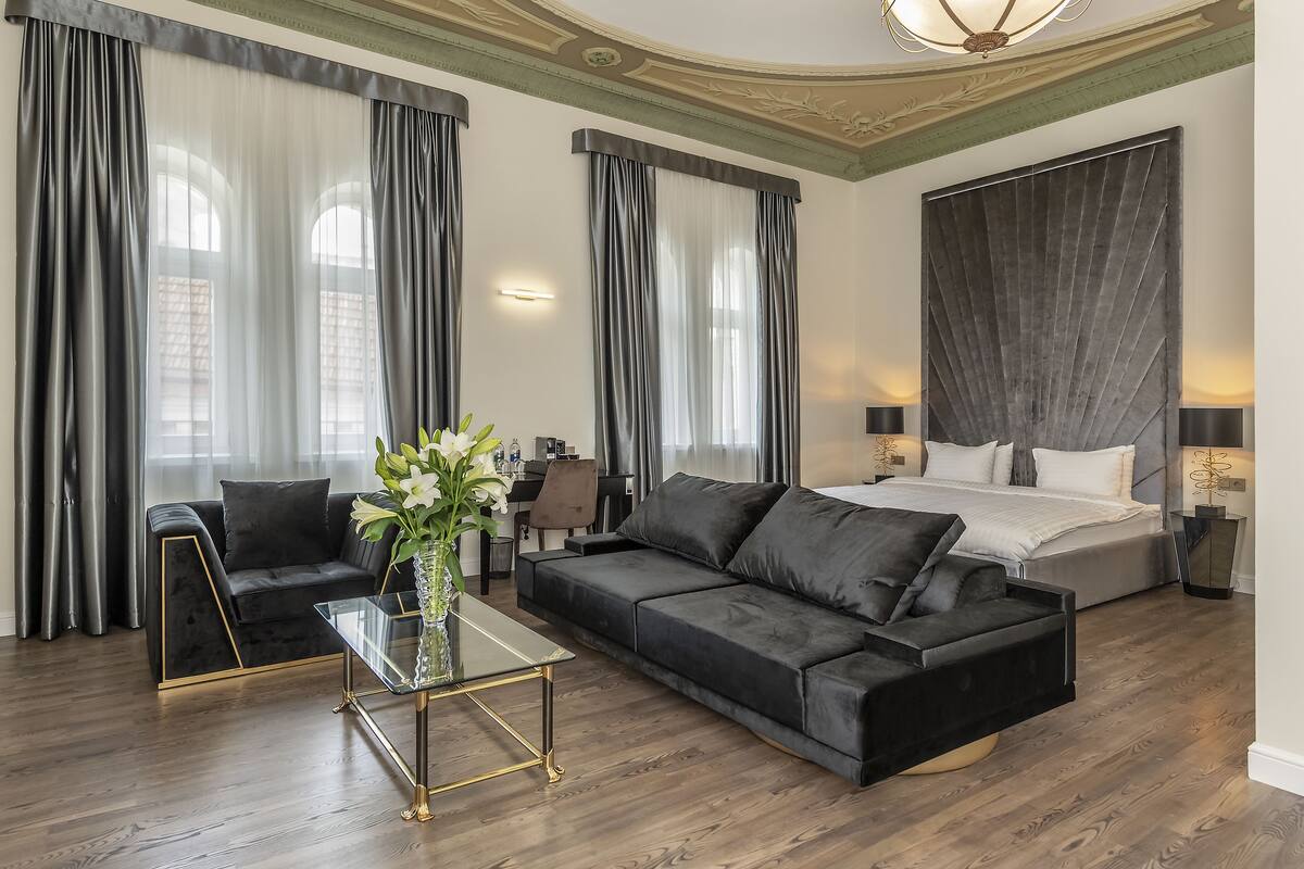 A modern hotel room with a black leather sofa, a glass coffee table, a bed with white linens, and large windows with sheer curtains.