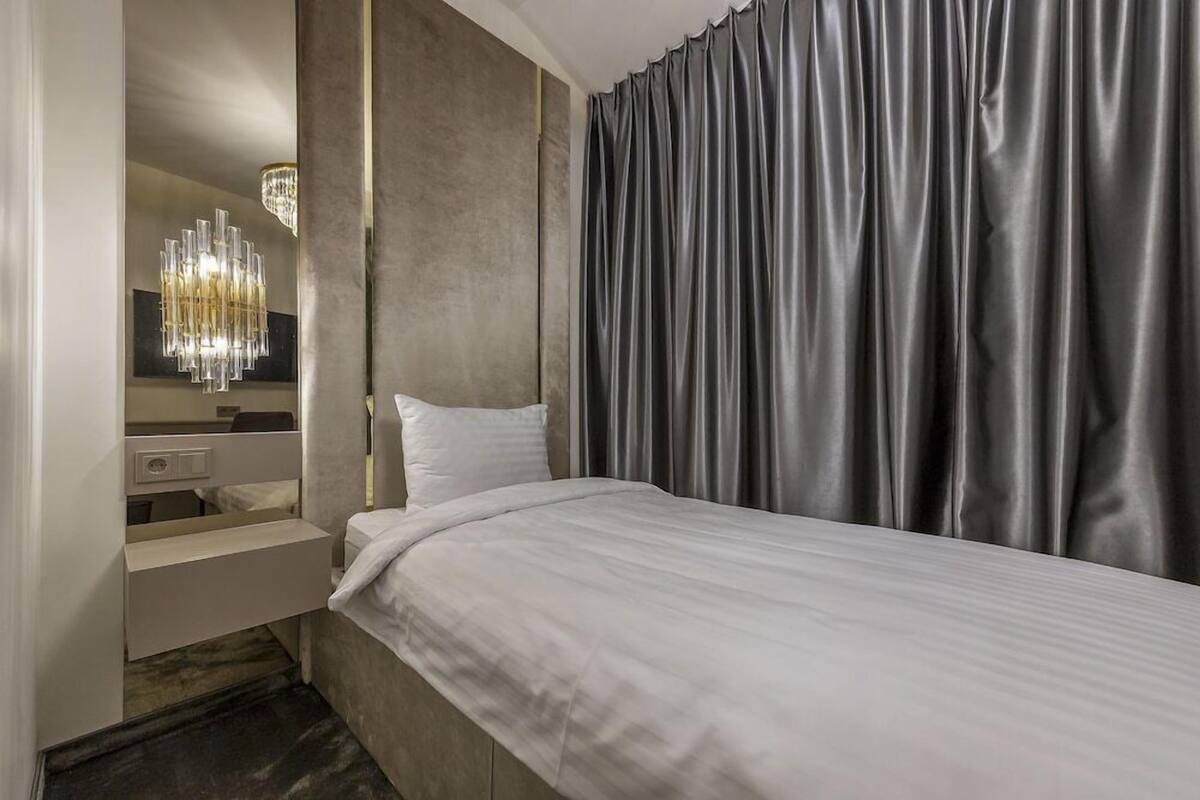 A hotel room with a bed, a mirror, a chandelier, and grey curtains.