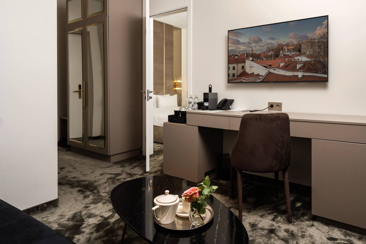 A hotel room with a desk, a chair, a coffee table with a teapot and a flower, a painting on the wall, and a mirror reflecting the room.