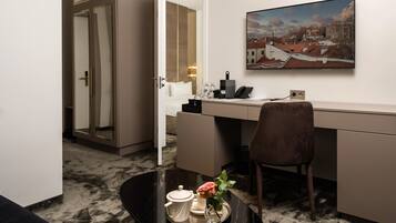 A hotel room with a desk, a chair, a coffee table with a teapot and a flower, a painting on the wall, and a mirror reflecting the room.