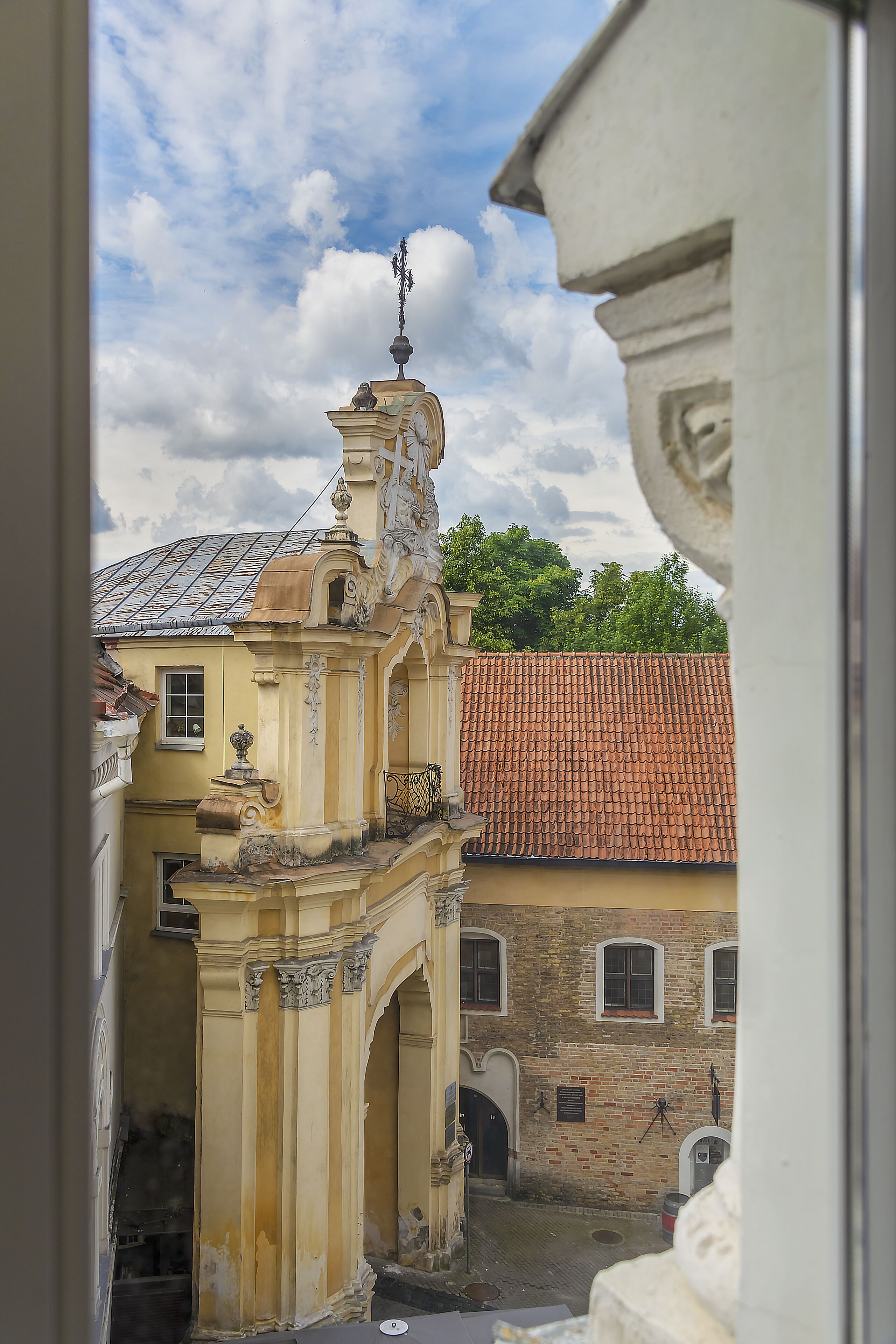 Vista de un patio con un edificio amarillo de arquitectura ornamentada, un techo de tejas rojas y una estatua en la parte superior.