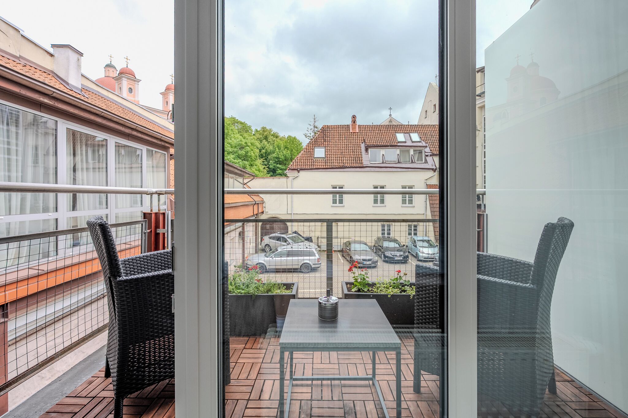 Een balkon met rieten stoelen, een tafel en uitzicht op geparkeerde auto's en gebouwen.
