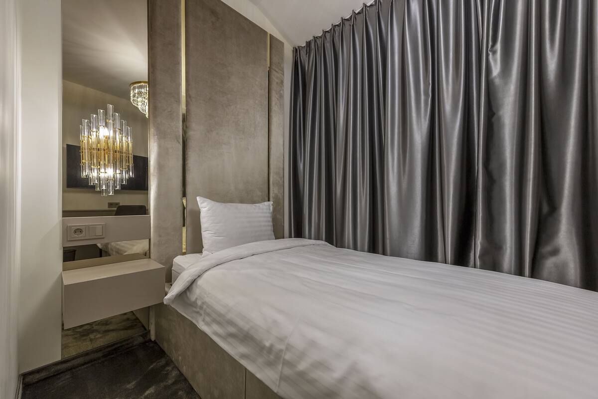 A modern hotel room with a bed, a vanity with a mirror, a chandelier, and grey curtains.