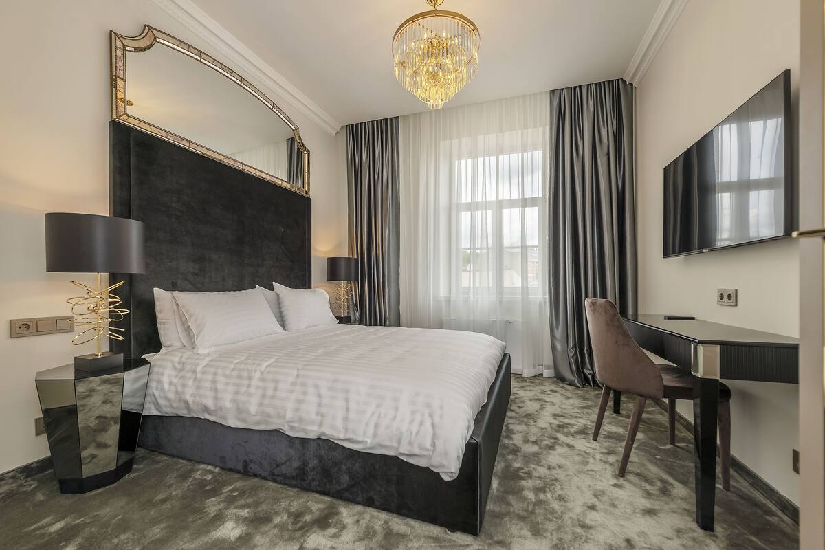 A modern hotel room with a large bed, a desk, a chair, a TV, and a chandelier.