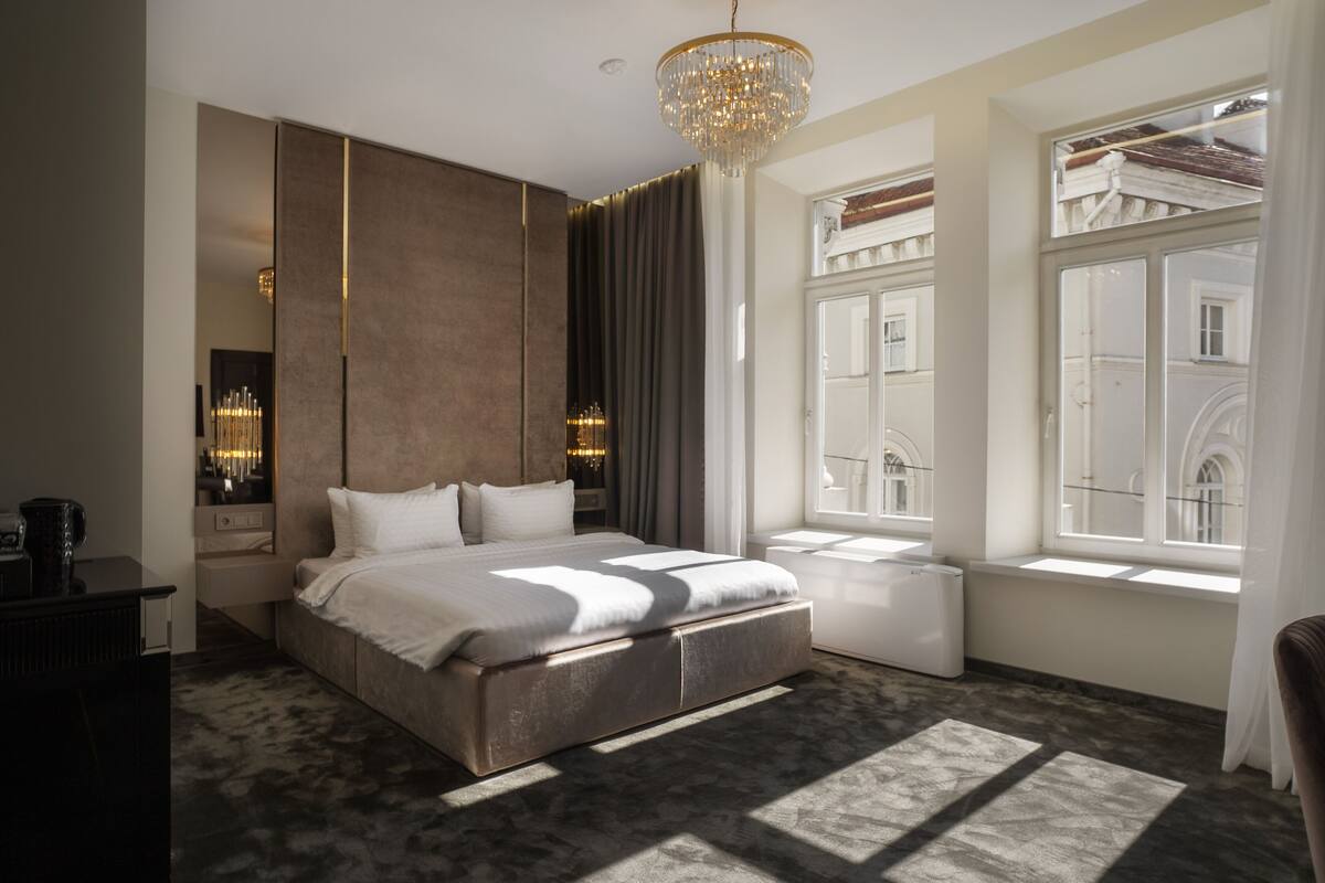 A modern bedroom with a large bed, a chandelier, a view of the city through large windows, and a small bedside table.