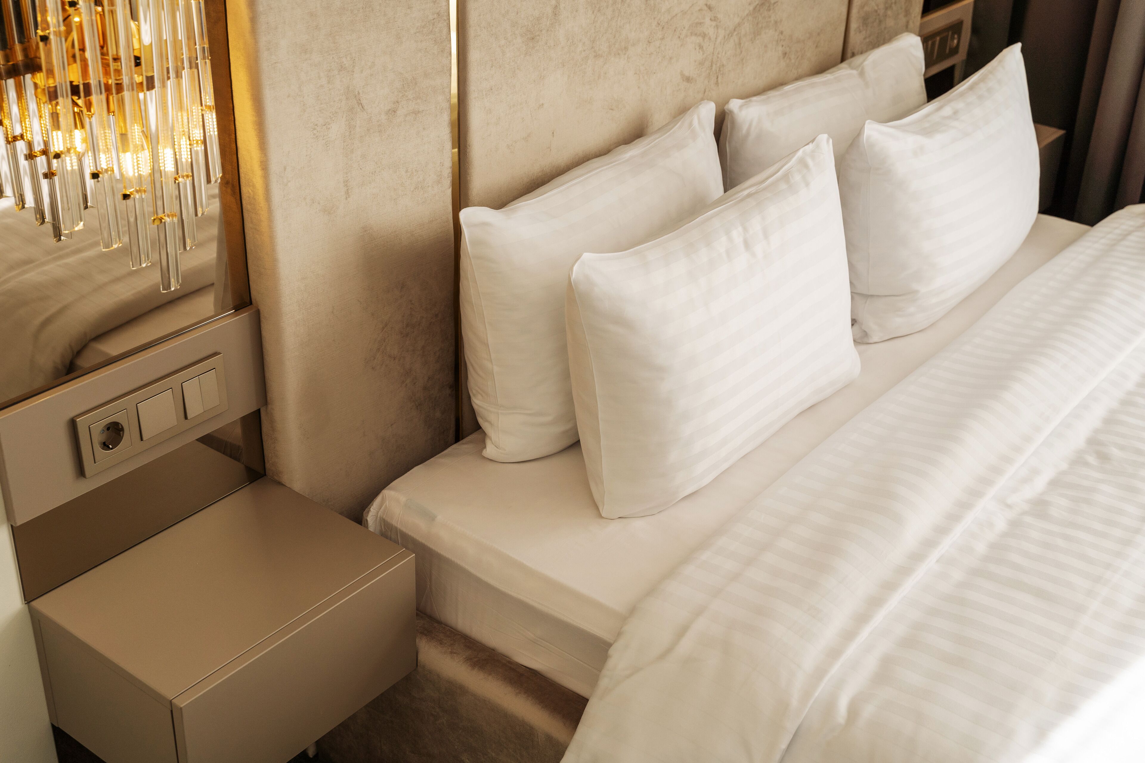 Een net opgemaakt bed met witte lakens en kussens, een nachtkastje met een lamp en een wandlamp.