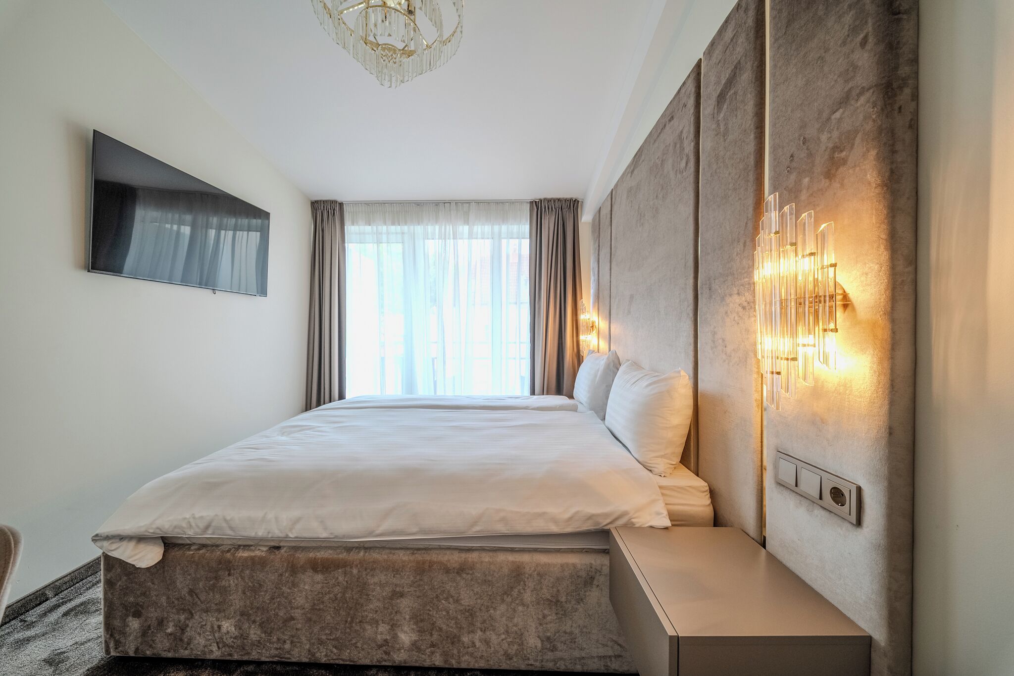 A modern hotel room with a large bed, a flat-screen TV, a wall-mounted light fixture, and a view of the city through large windows.