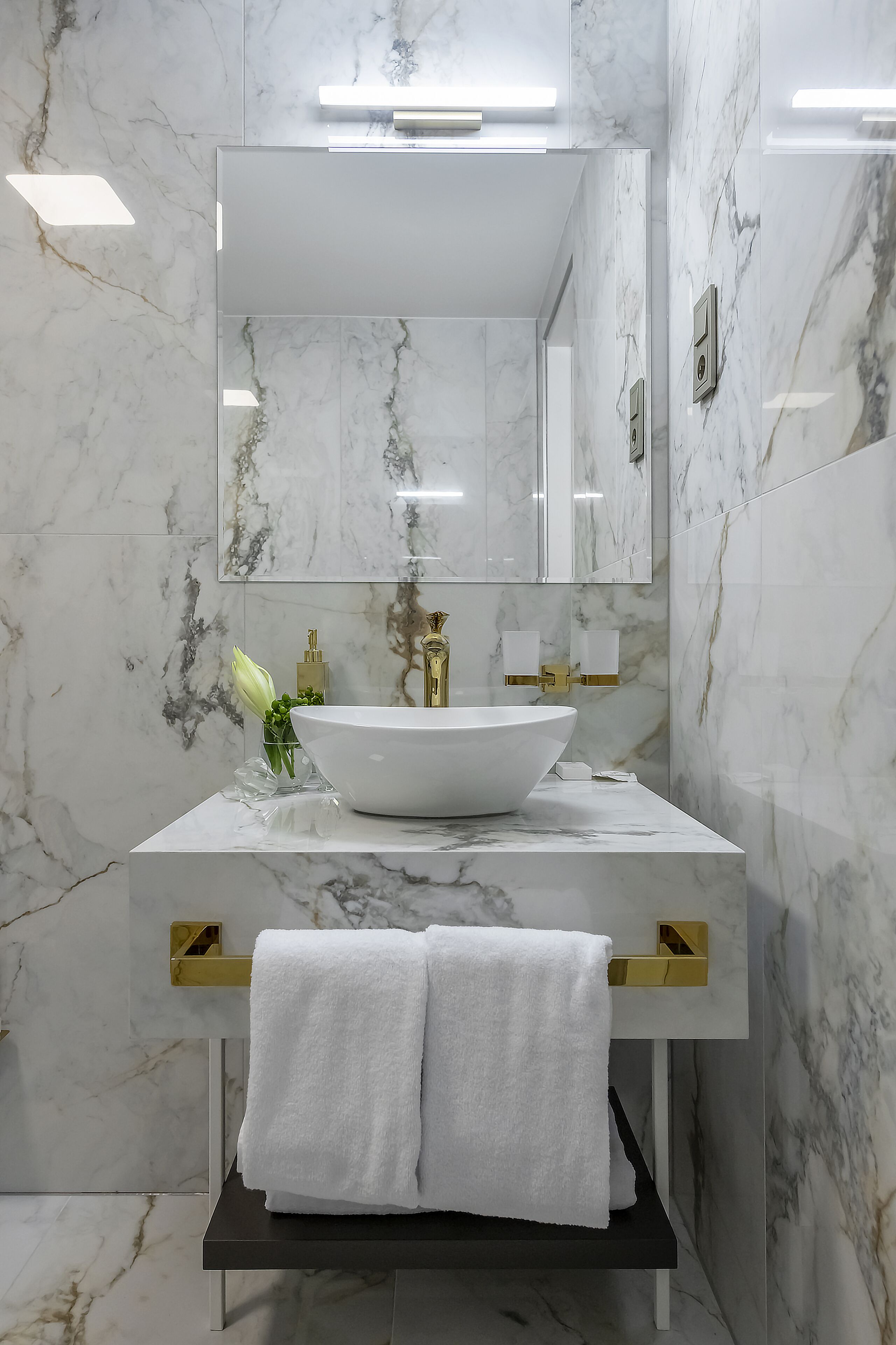A modern bathroom with a marble backsplash, a white sink, gold fixtures, and a large mirror.