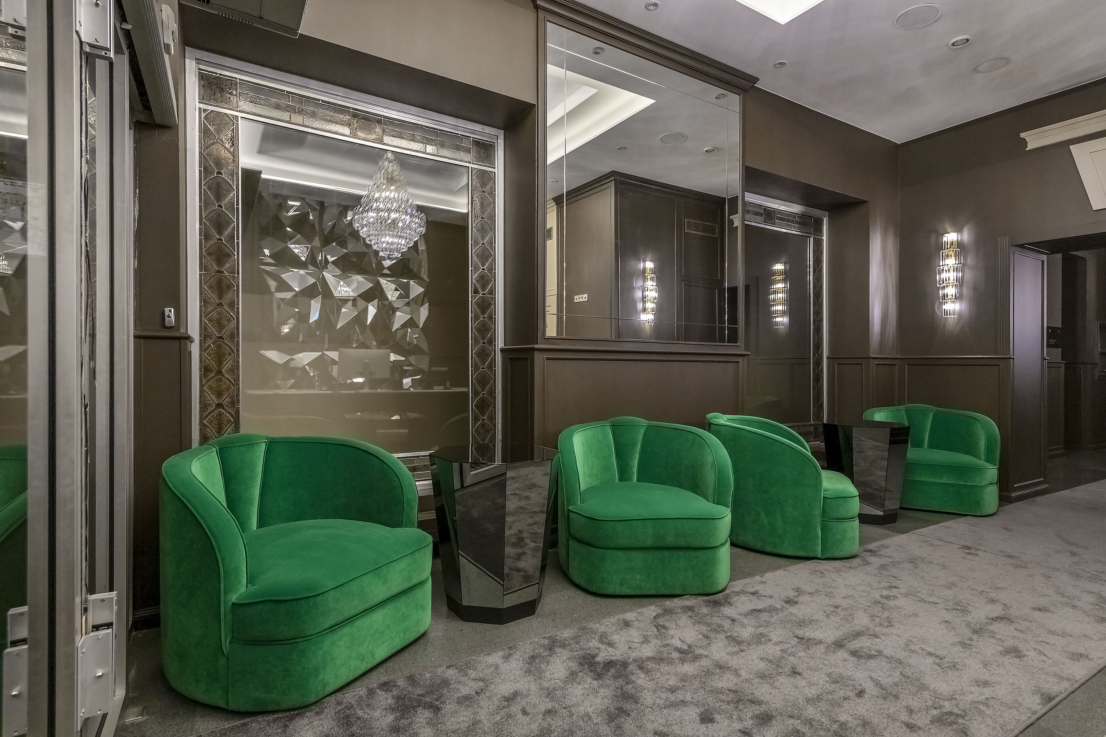 A modern hotel lobby with green upholstered chairs, a large mirror, and a chandelier.
