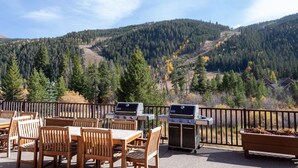 An outdoor patio with wooden tables and chairs, barbecue grills, and a view of a forested mountain.