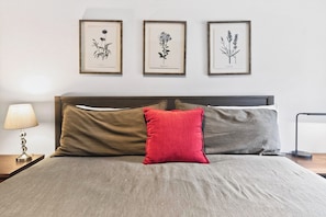 A bed with a grey and brown bedspread, two pillows, a nightstand with a lamp, and three framed botanical illustrations on the wall.