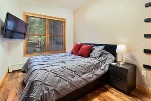 Une chambre à coucher avec un lit, une table de chevet avec une lampe, un téléviseur fixé au mur et une fenêtre avec des stores.