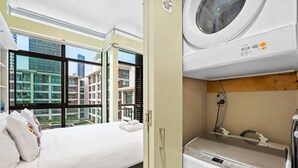 Una camera d'albergo con un letto, una lavatrice e vista sugli edifici attraverso ampie finestre.