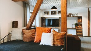 Un salon moderne avec un canapé noir, des poutres en bois, un téléviseur et une bibliothèque.