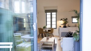 Una sala moderna con sofá, una mesa de centro y un comedor con sillas y una mesa. Se observa una zona de cocina con ventana y una planta.