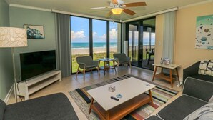 A modern living room with a TV, seating, a coffee table, and a balcony with a view of the beach.