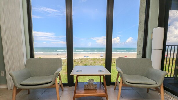 A room with a view of the beach, two grey chairs, a small table, and a window with a view of the ocean.