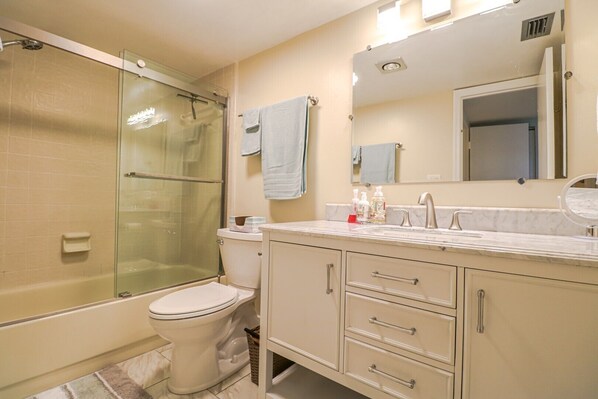 A bathroom with a glass-enclosed shower, a toilet, a sink with a marble countertop, and a mirror. - Sand Dollar 2-202 - Beautiful Newly Remodeled Oceanfront unit on the 2nd floor at Sand Dollar! (St. Augustine)