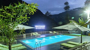 A swimming pool with lounge chairs and umbrellas, surrounded by greenery and a mountainous backdrop. - Bhotekoshi Heli Resort and Spa (Marming)