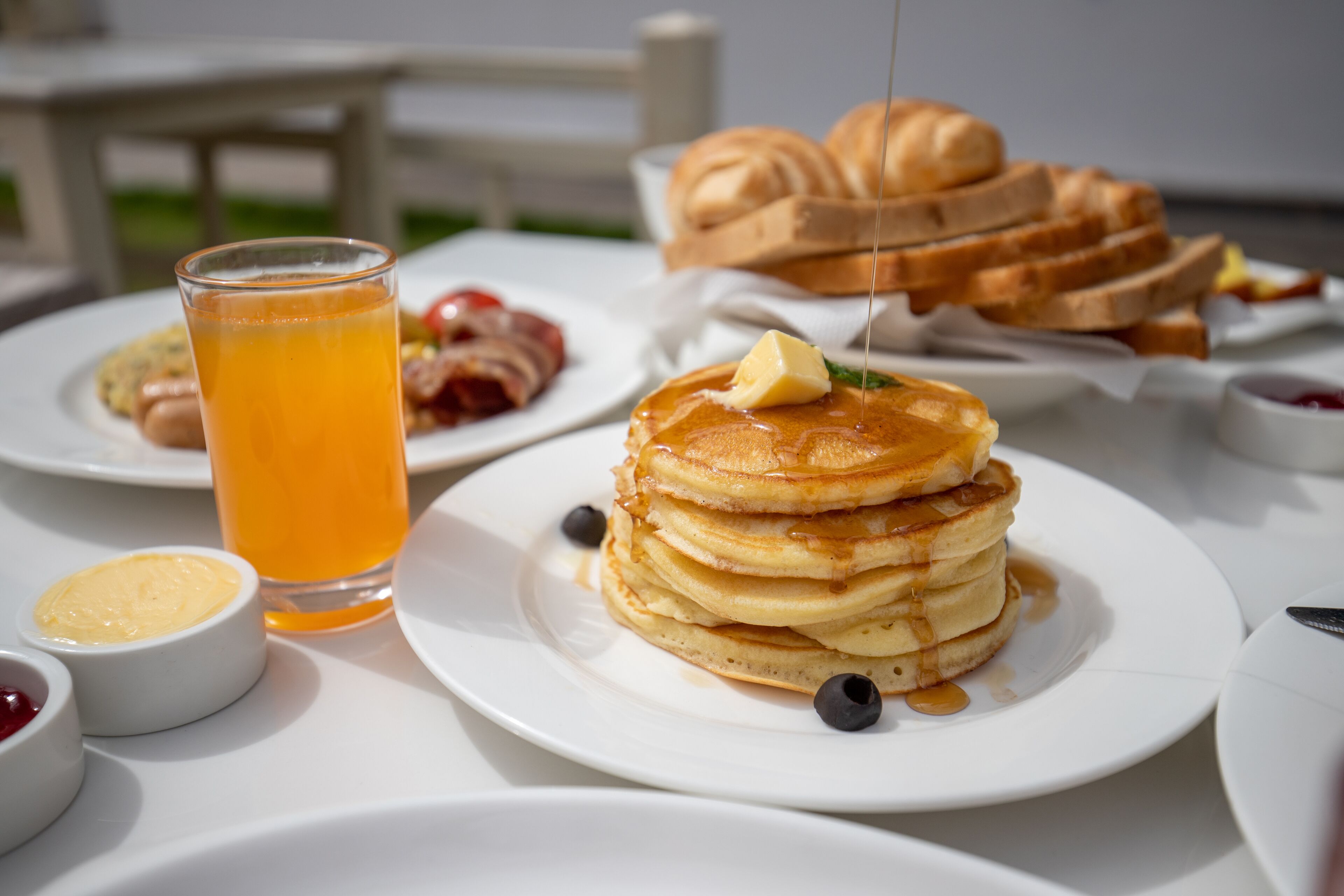 Desayuno con tortitas, tostadas y un vaso de zumo de naranja sobre una mesa blanca.