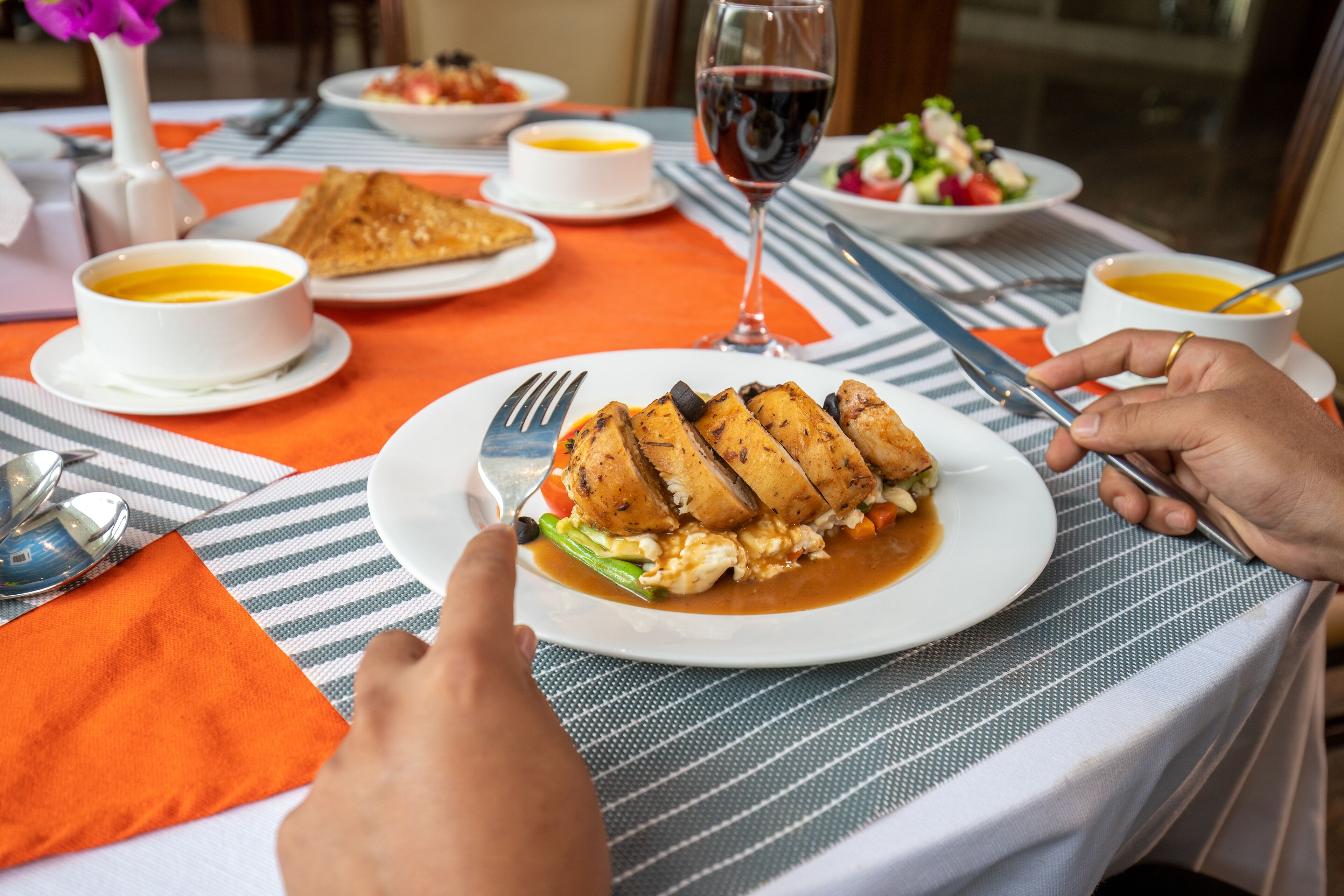 A dining table with a plate of grilled chicken, vegetables, and sauce, accompanied by bowls of soup and a glass of red wine.