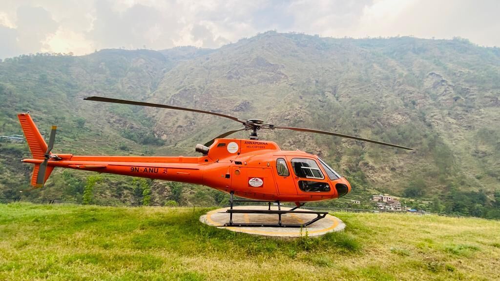 A red helicopter with "ANNAPURNA HELICOPTERS" and "9N-ANU 3" on its side, parked on a grassy area with a mountainous backdrop.