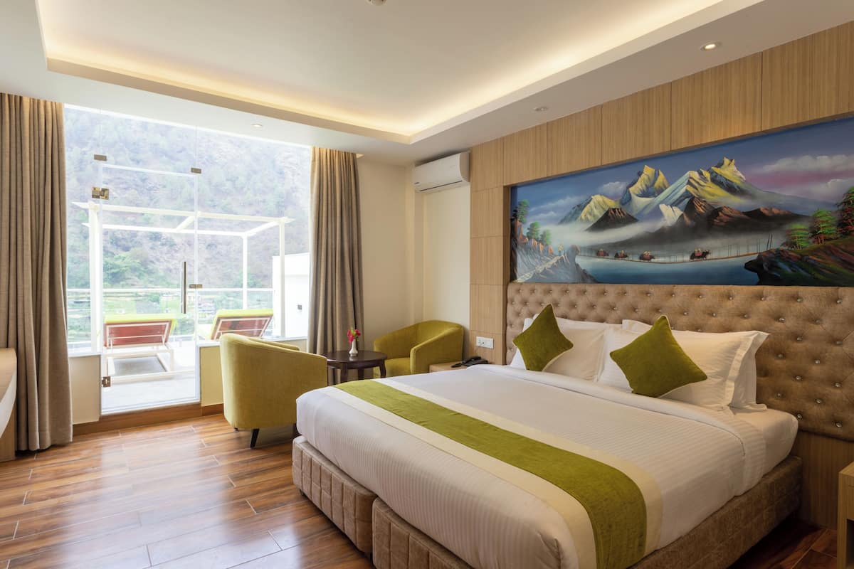 Habitación de hotel con una cama grande, un cuadro de montañas, un balcón con sillas y vistas a zonas verdes.