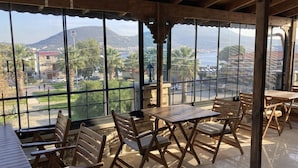 A covered outdoor seating area with wooden tables and chairs, large windows offering a view of a town and mountains. - Ionia Liberty Stone Hotel (Foça)