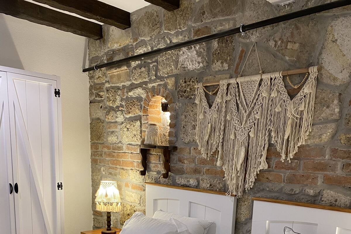A bedroom with two beds, a stone wall, a macrame wall hanging, and a nightstand with a lamp.