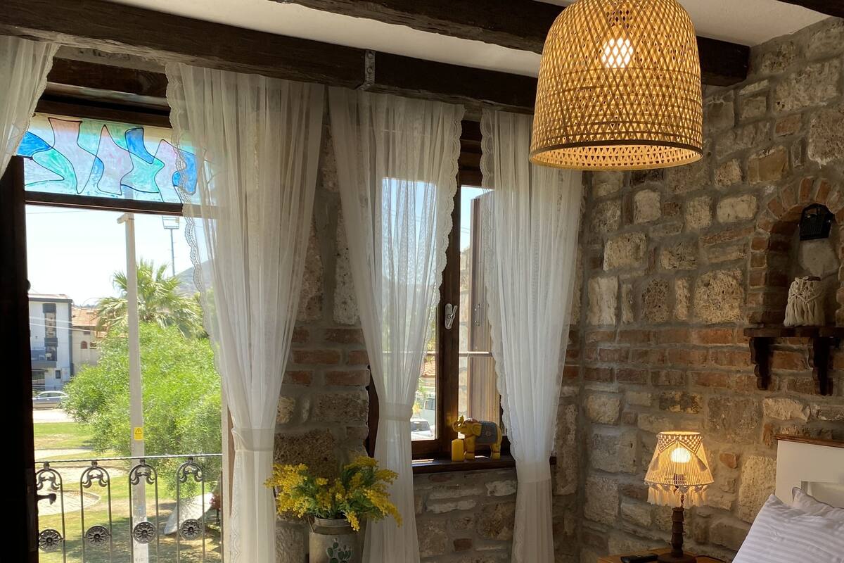 A bedroom with a large bed, a hanging light fixture, a nightstand with a lamp, and a view of the outside balcony.