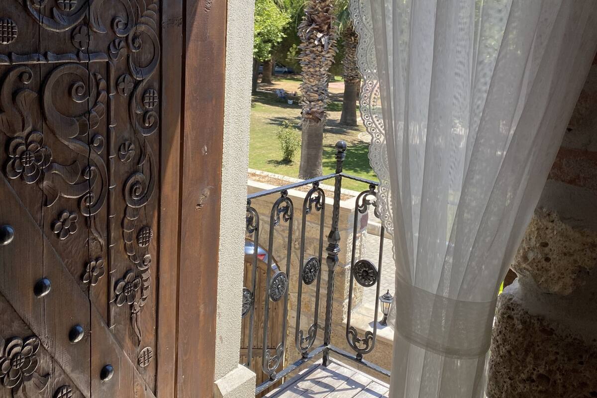 A wooden door with intricate carvings, a balcony with a wrought iron railing, and a view of a garden with palm trees.