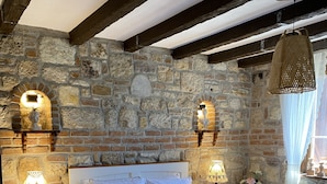 A bedroom with a stone wall, a wooden ceiling, a bed with white bedding, two bedside tables with lamps, and a window with sheer curtains. - Ionia Liberty Stone Hotel (Foça)