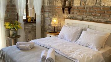 Un dormitorio con pared de piedra, una cama con ropa blanca, una lámpara colgante y una mesita de noche con lámpara.