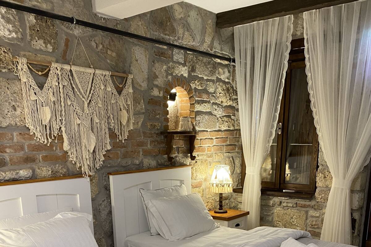 A bedroom with a stone wall, a bed with white bedding, a nightstand with a lamp, and a window with curtains.