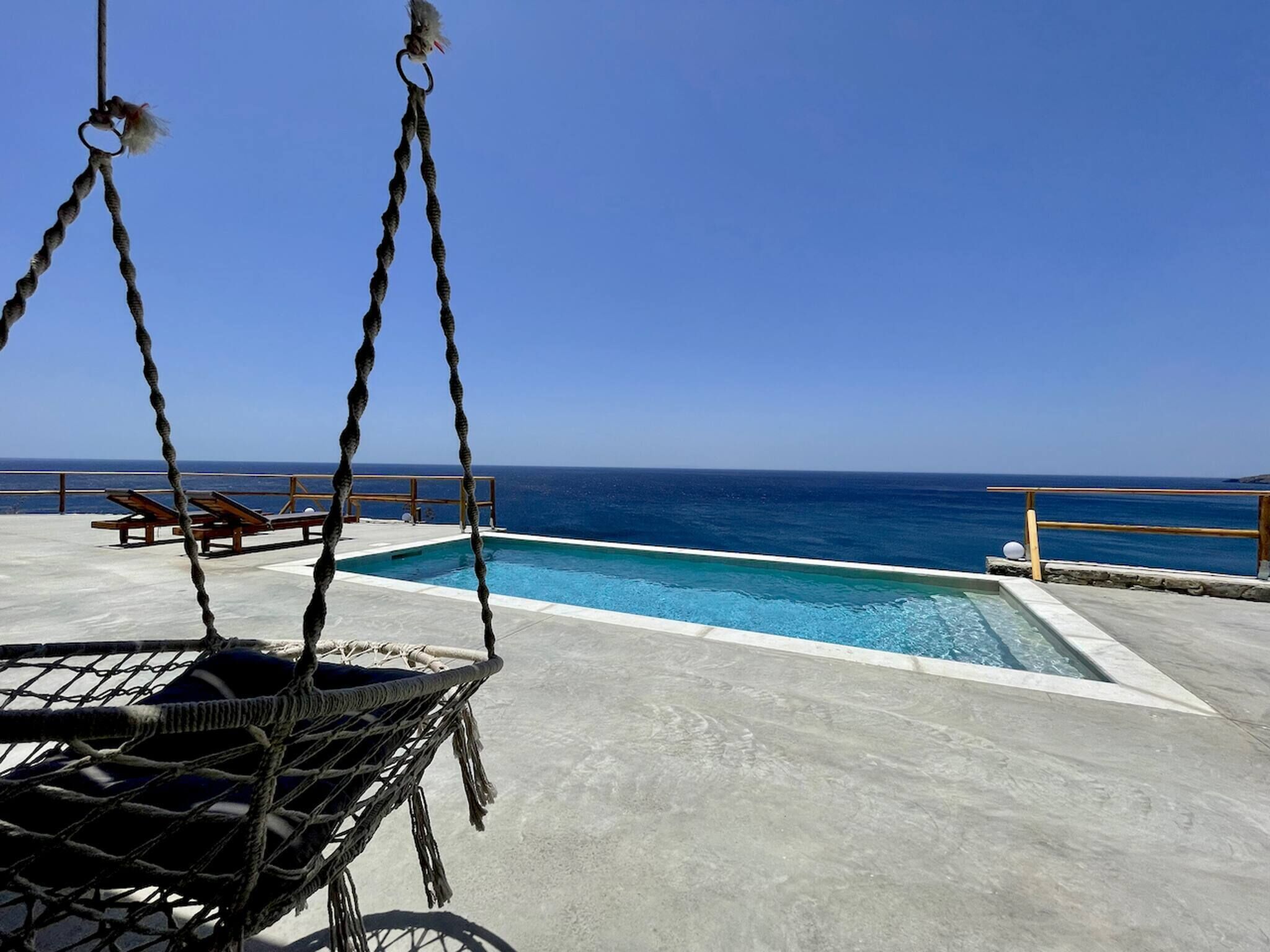 Une piscine avec vue sur l’océan, un fauteuil suspendu et des chaises longues en bois.