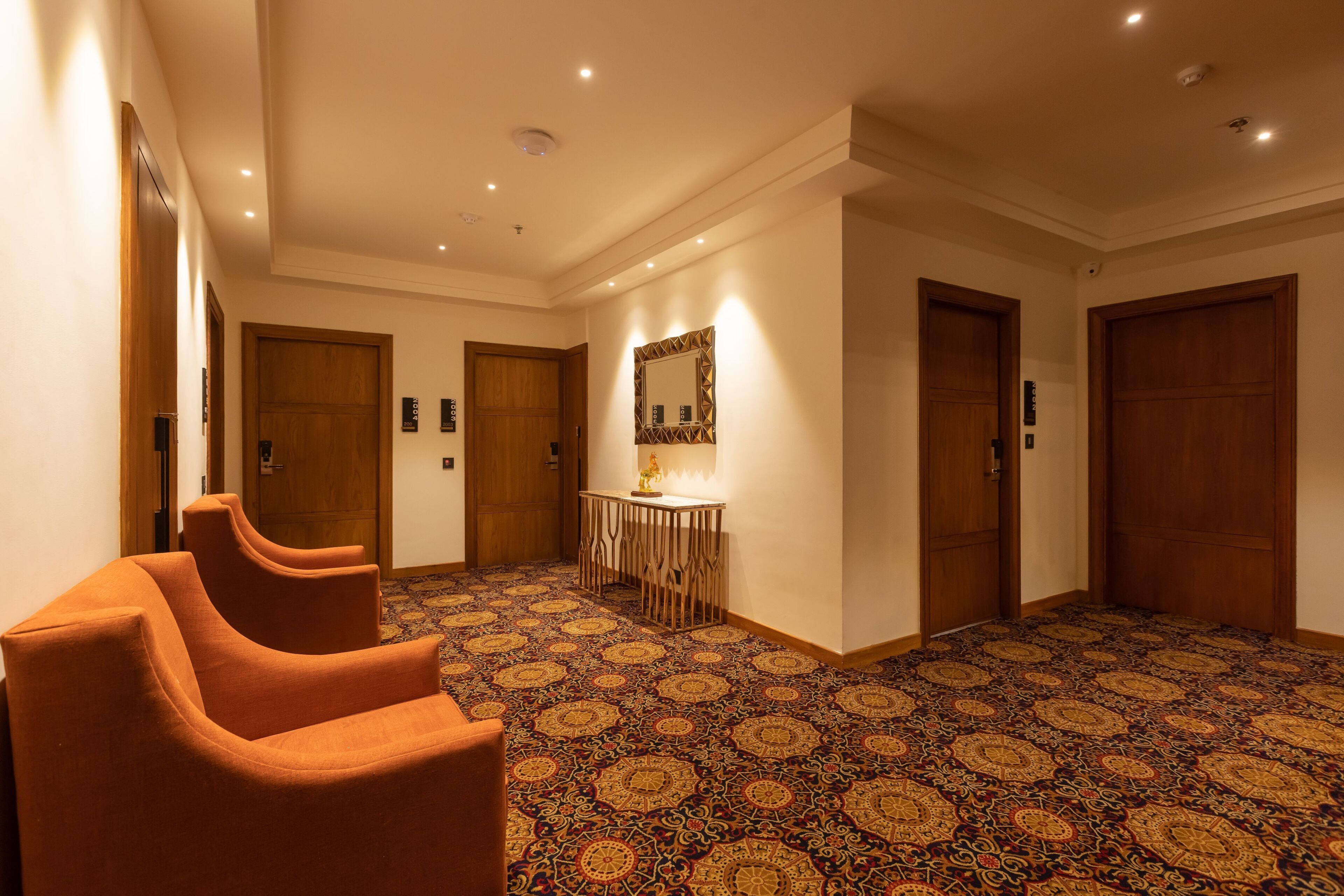 Ein Hotelgang mit gemustertem Teppich, Holztüren und einem Tisch mit Lampe.