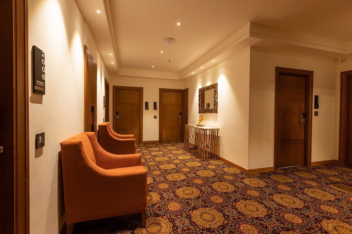 A hotel hallway with a patterned carpet, wooden doors, and a seating area with orange chairs.