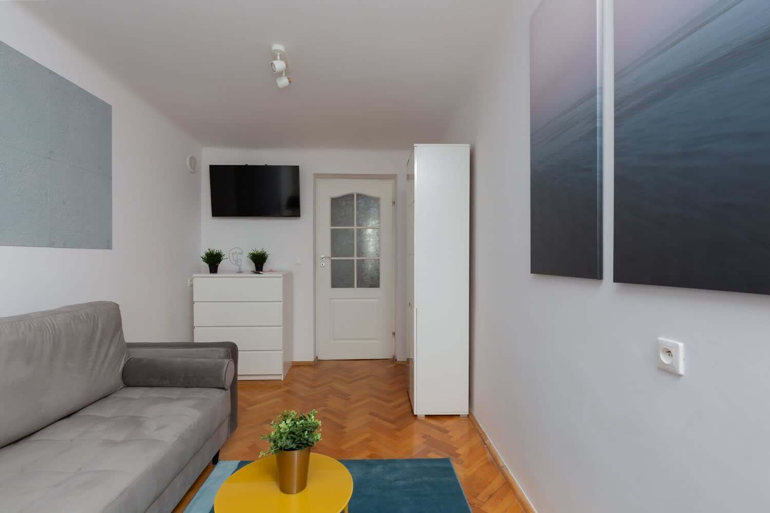 Una sala de estar moderna con un sofá gris, un aparador blanco, un televisor de pantalla plana y una puerta que da a otra habitación.