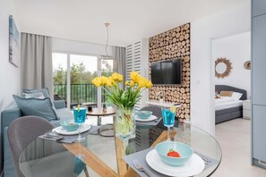 A modern living room with a glass dining table, a sofa, a TV mounted on a wooden wall, and a balcony view.