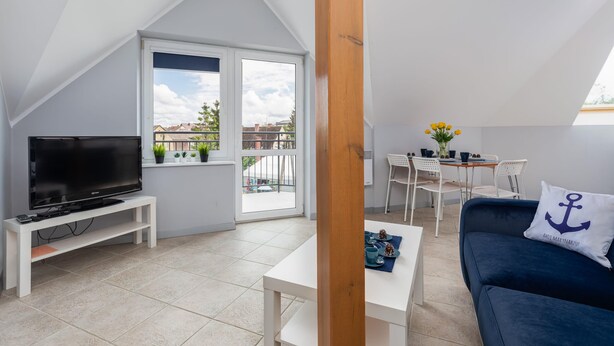 A modern living room with a TV on a stand, a dining area with a table and chairs, and a balcony with plants. - Family Apartment for 6 Persons | Close to the Beach and Parking (Pobierowo)