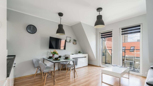 Una cocina moderna con comedor, un televisor montado en la pared y un balcón con vistas a edificios de tejados rojos.